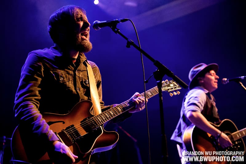 Two members of The Wild Feathers singing and playing guitars under blue stage light at the 9:30 Club in Washington, D.C.
