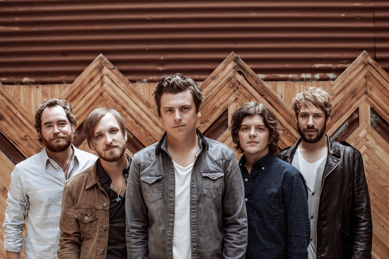 Five members of The Wild Feathers posed shoulder to shoulder before a chevron-patterned wooden wall.