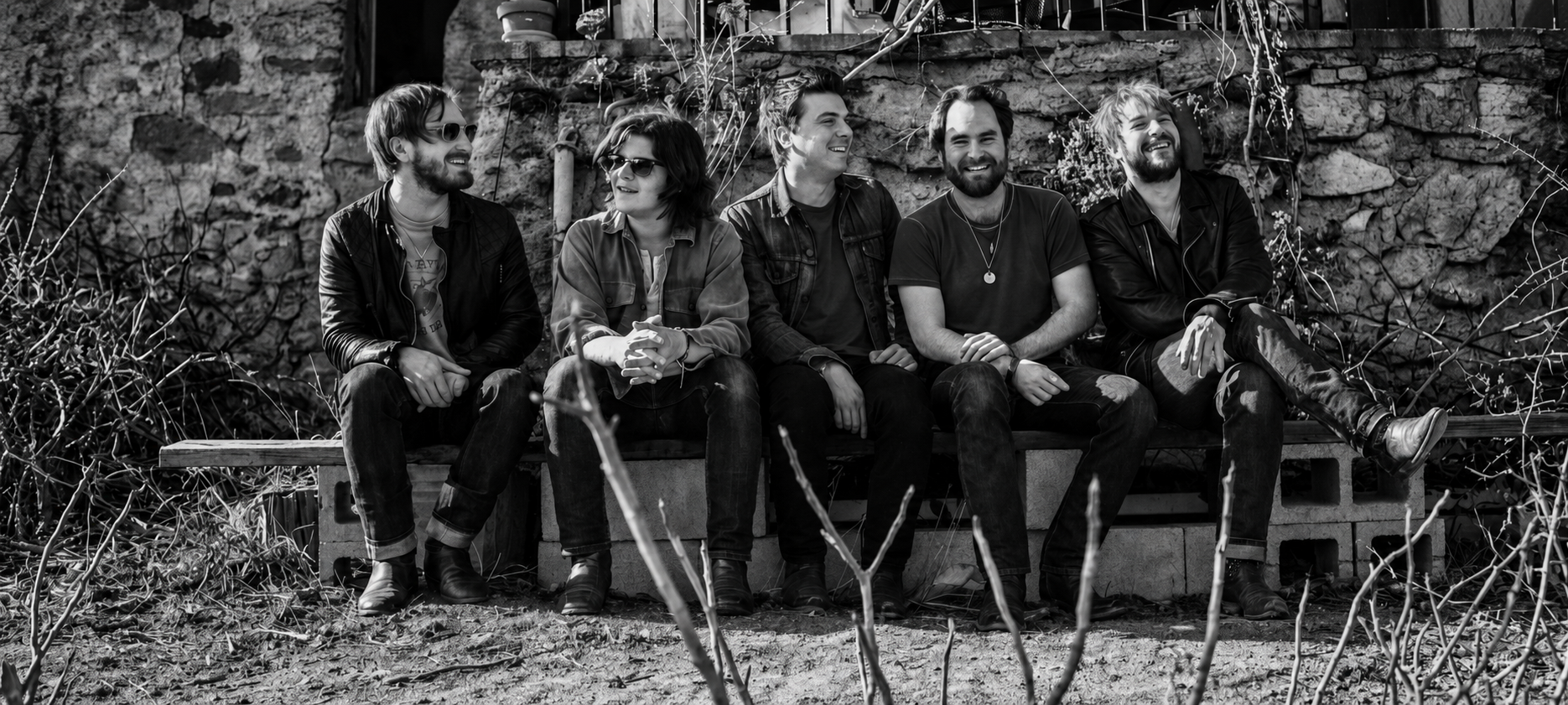 Five members of The Wild Feathers seated together outdoors in front of a rough stone wall, smiling and looking toward one another in a black-and-white panoramic portrait.