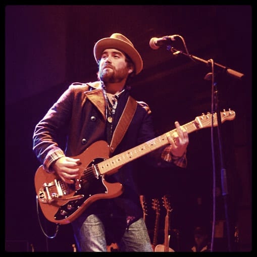 A square live photograph of a Wild Feathers guitarist performing under concert lighting.