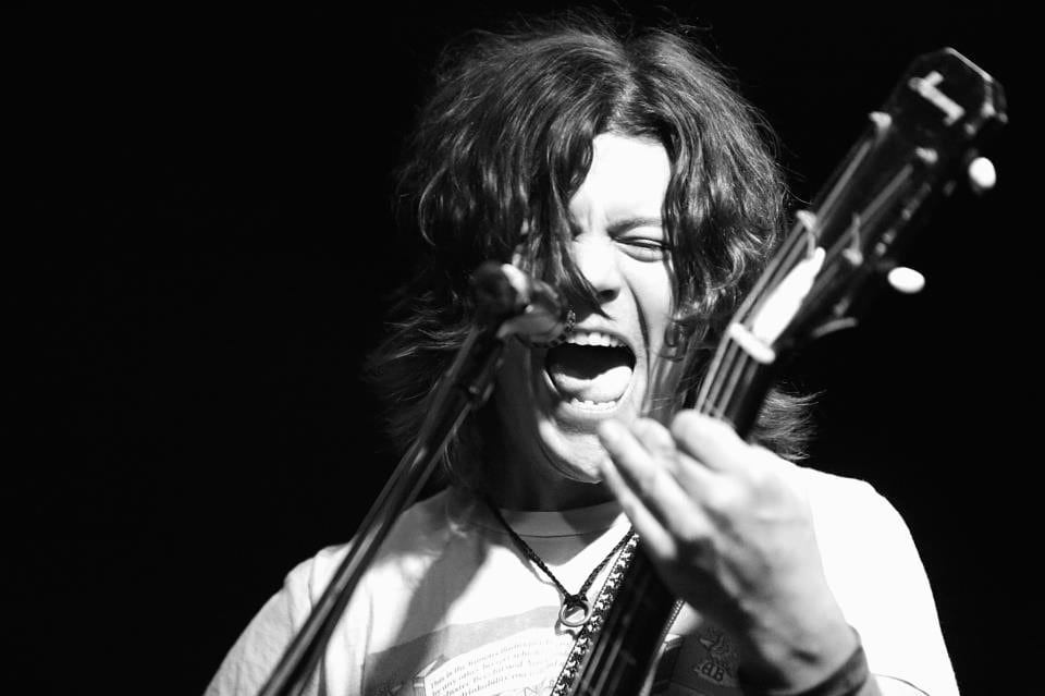 A professional black-and-white close view of a Wild Feathers guitarist singing onstage.