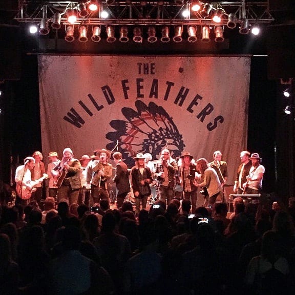 Crowded concert stage beneath a large Wild Feathers backdrop with the audience visible in the foreground.