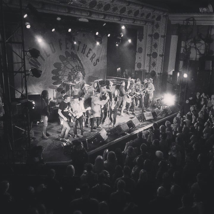 Black-and-white elevated view of a crowded theater stage beneath a Wild Feathers backdrop with the audience below.