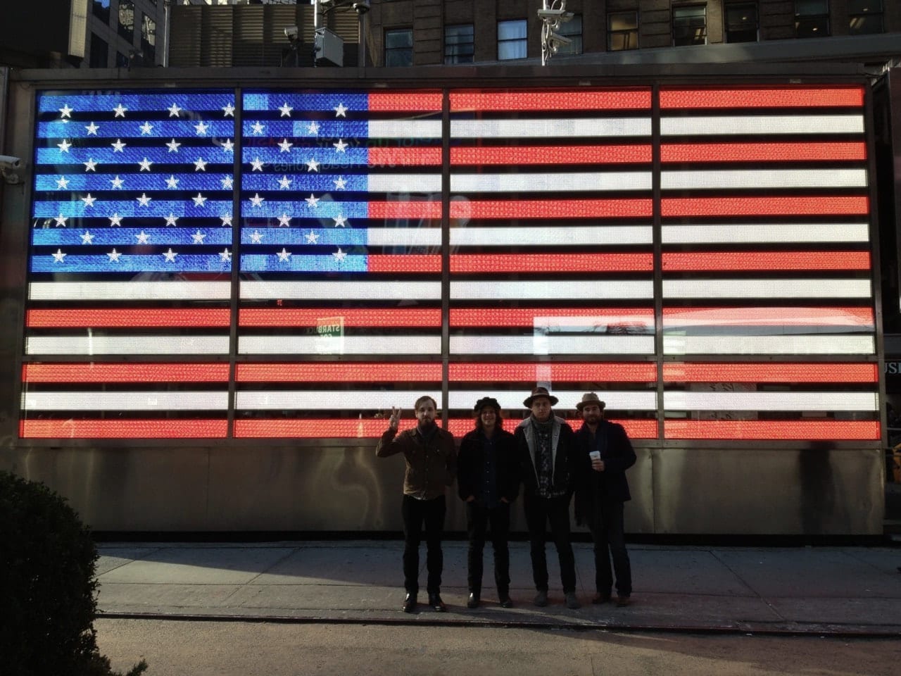 Four members of The Wild Feathers standing in front of the illuminated American-flag facade of the U.S. Armed Forces Recruiting Station in Times Square.