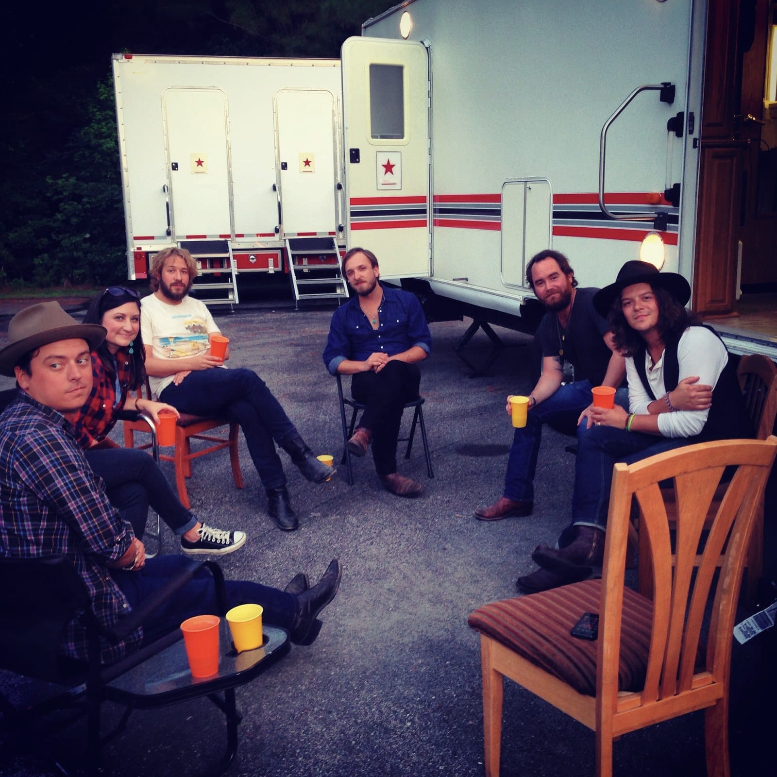 Five people sit outdoors on mismatched chairs beside two trailers during a travel day.
